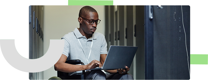 A man in a wheelchair wearing glasses, a gray polo shirt, and a lanyard is working on a laptop in a server room. He has a focused expression as he types, surrounded by tall, dark server racks. The environment is well-lit, with a contrast between the bright hallway and the darker server cabinets.